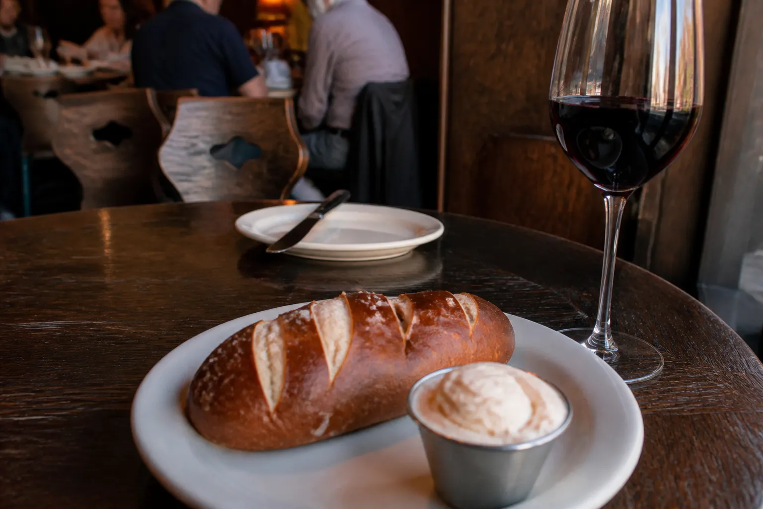 A golden-brown pretzel bread loaf with whipped butter and a glass of red wine on a restaurant table.