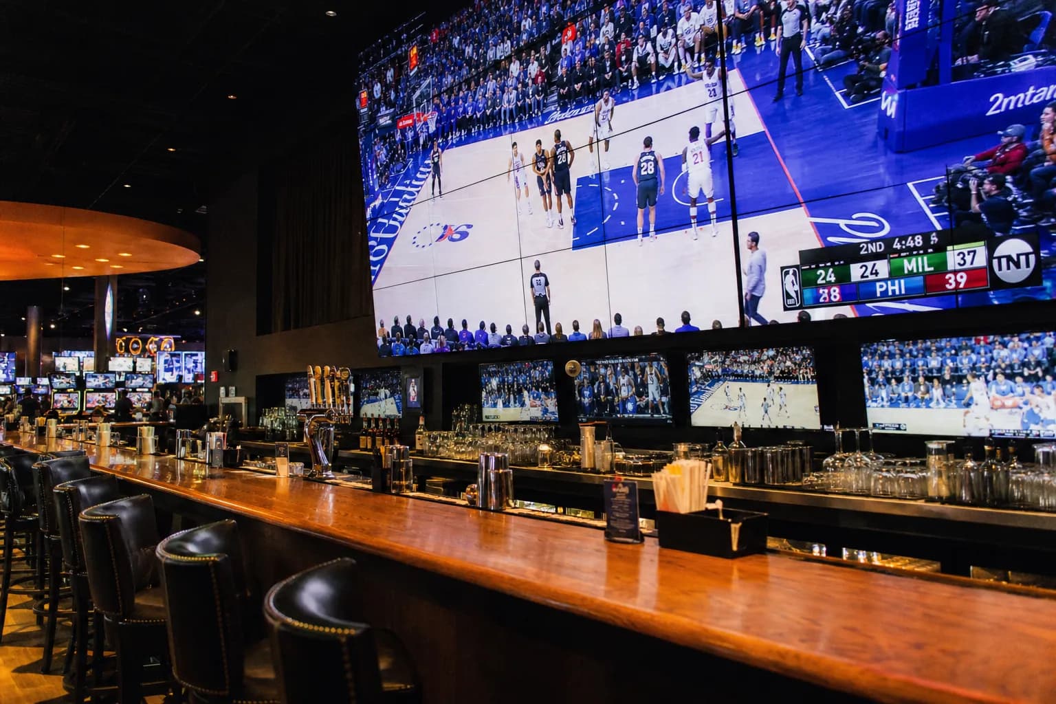 A vibrant, modern sports bar interior featuring a wide polished wooden counter, glowing neon overhead lights, and massive multi-screen LED displays showing live sports games.