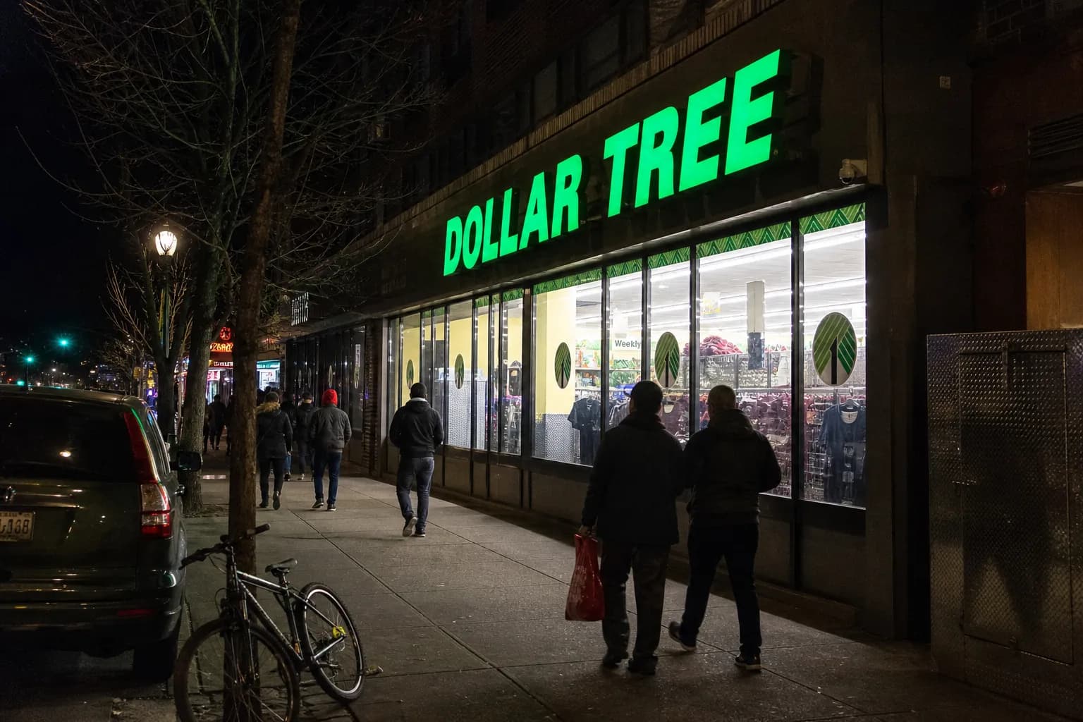 The brightly lit storefront of a Dollar Tree on Steinway Street in Astoria, Queens at night, with streetlights reflecting off the sidewalk.