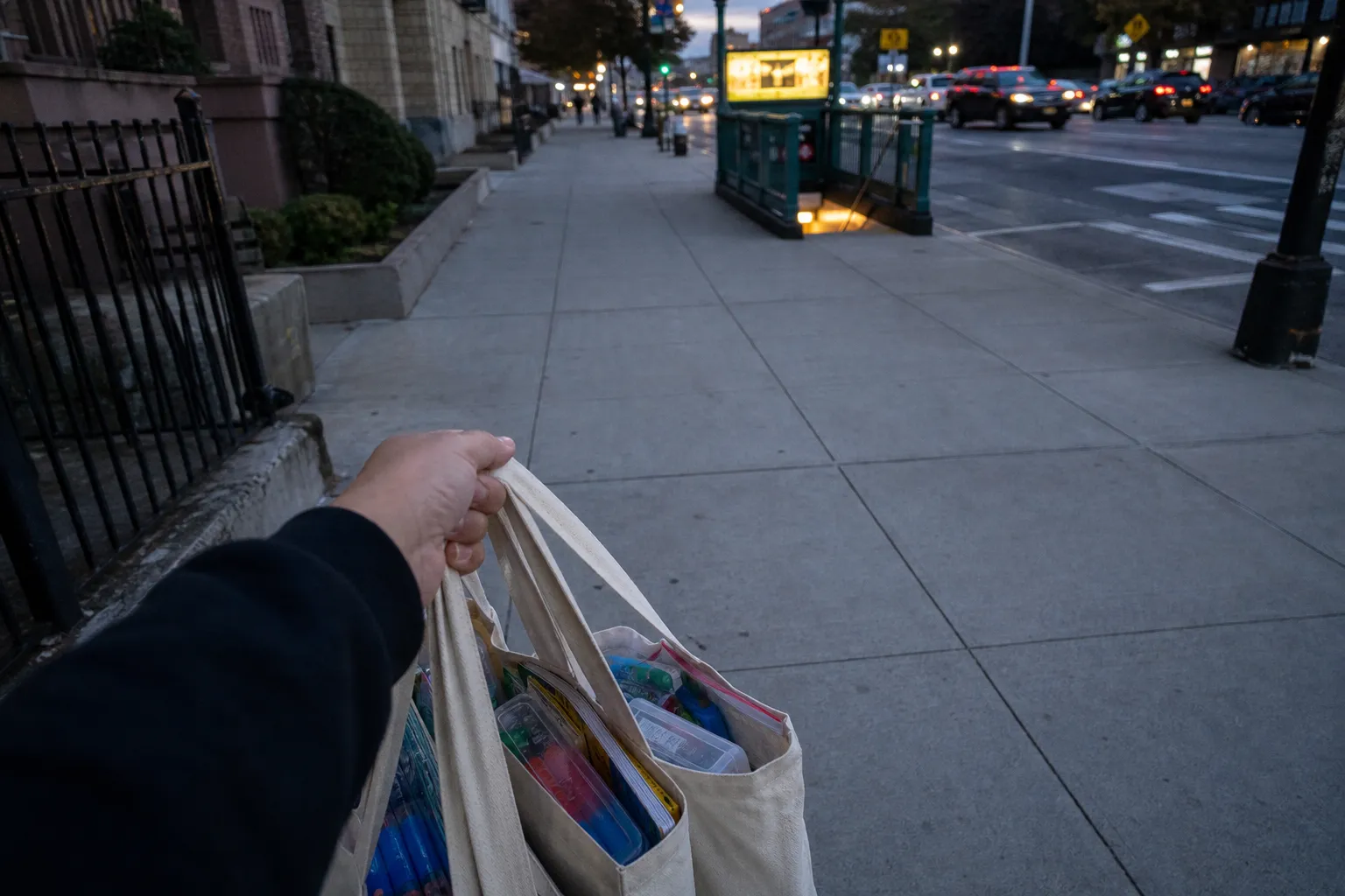 Shopper carrying loaded grocery bags near a Queens subway station entrance at sunset.