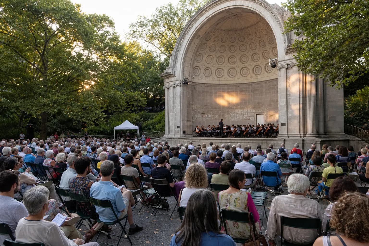 People sitting in folding chairs watching a classical orchestra perform at the Naumburg Bandshell at sunset.