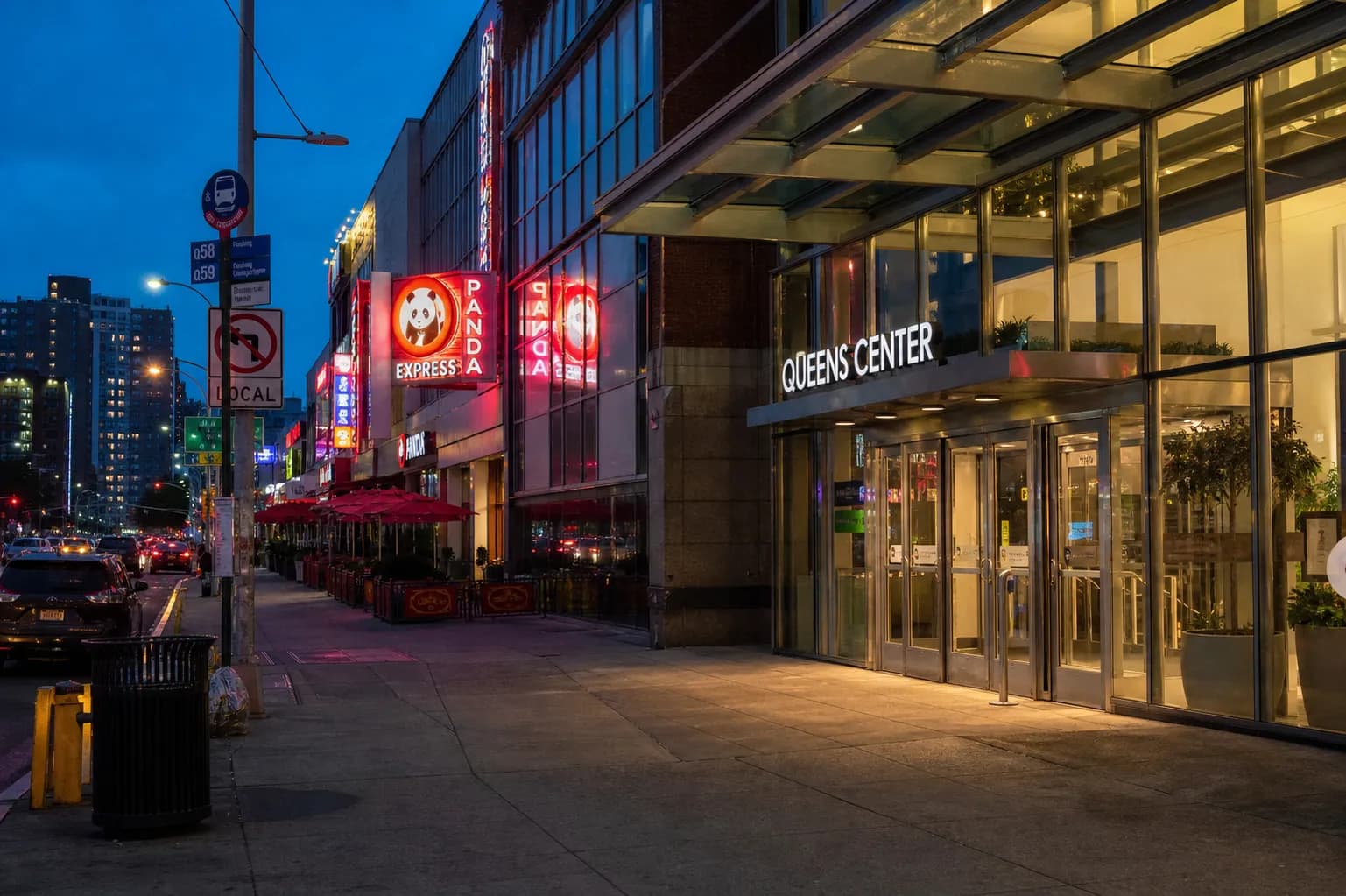 The busy sidewalk and glowing exterior storefronts outside Queens Center Mall at twilight.