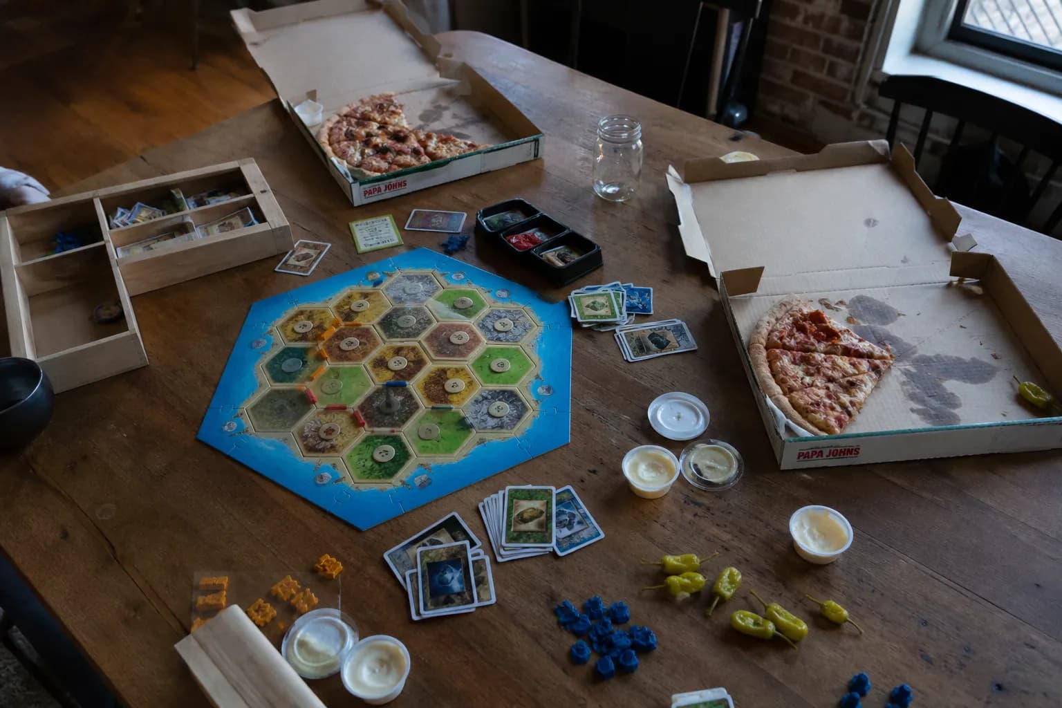 A casual tabletop setup during a board game night in a Brooklyn apartment, featuring open Papa Johns pizza boxes, garlic dipping sauces, and local craft beer cans.