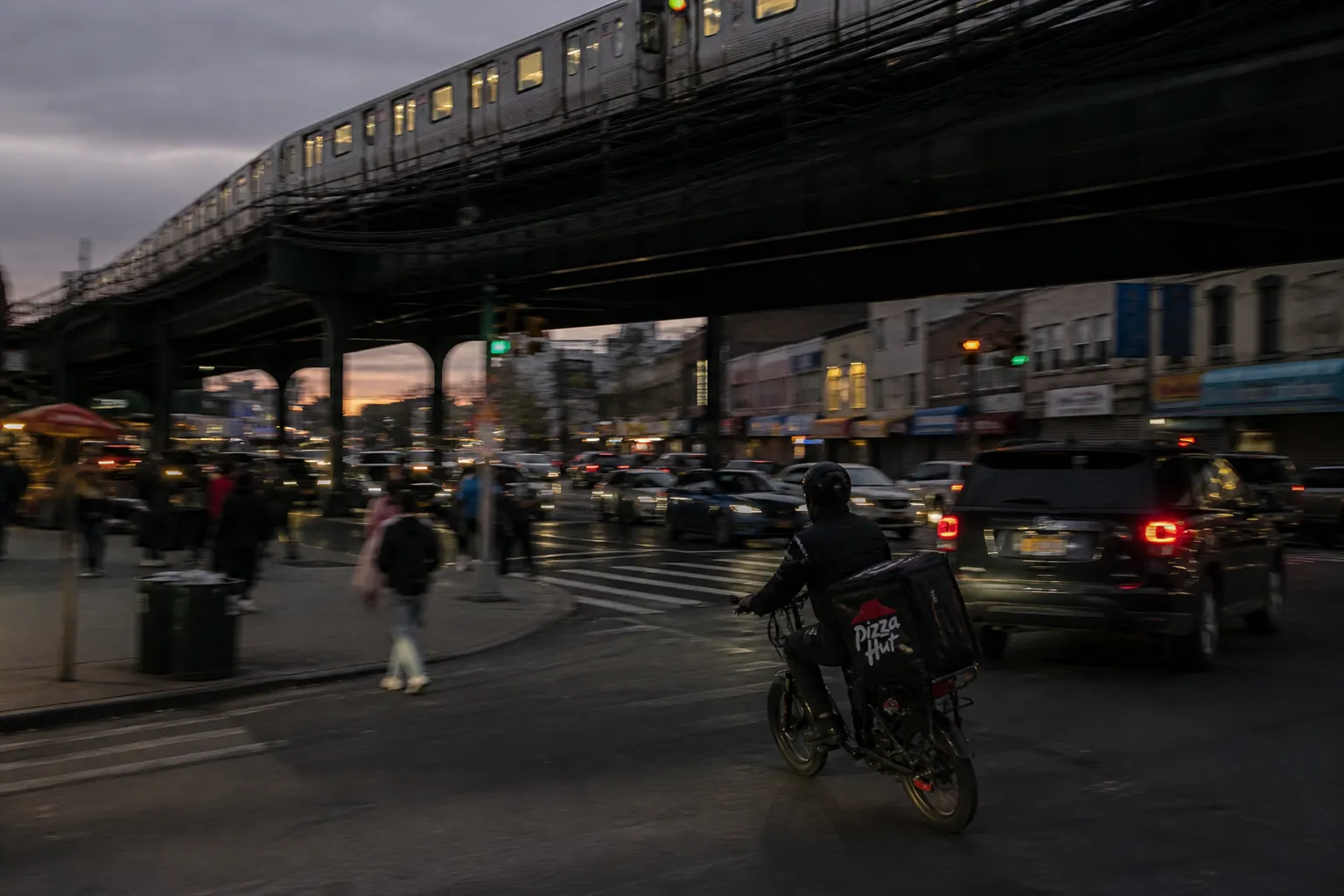 A delivery driver on a bicycle navigating a busy Queens street with the elevated subway line in the background.