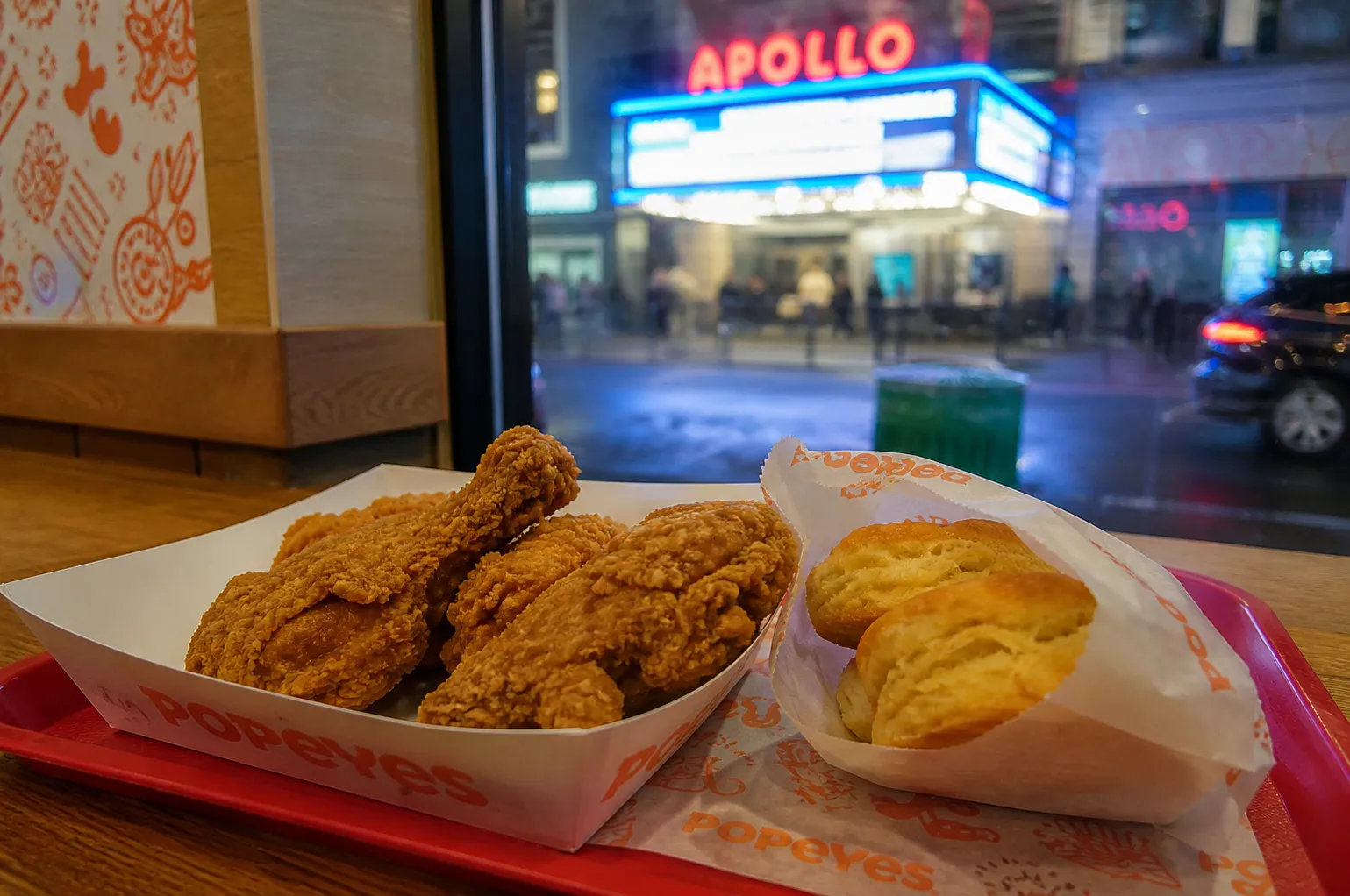 A glowing orange and red neon Popeyes sign reflecting on a wet Harlem sidewalk at night.