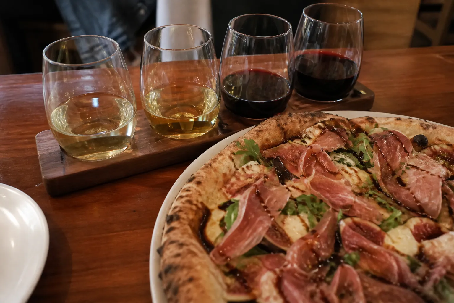 Four small wine glasses in a flight next to a freshly baked wood-fired pizza on a wooden table.