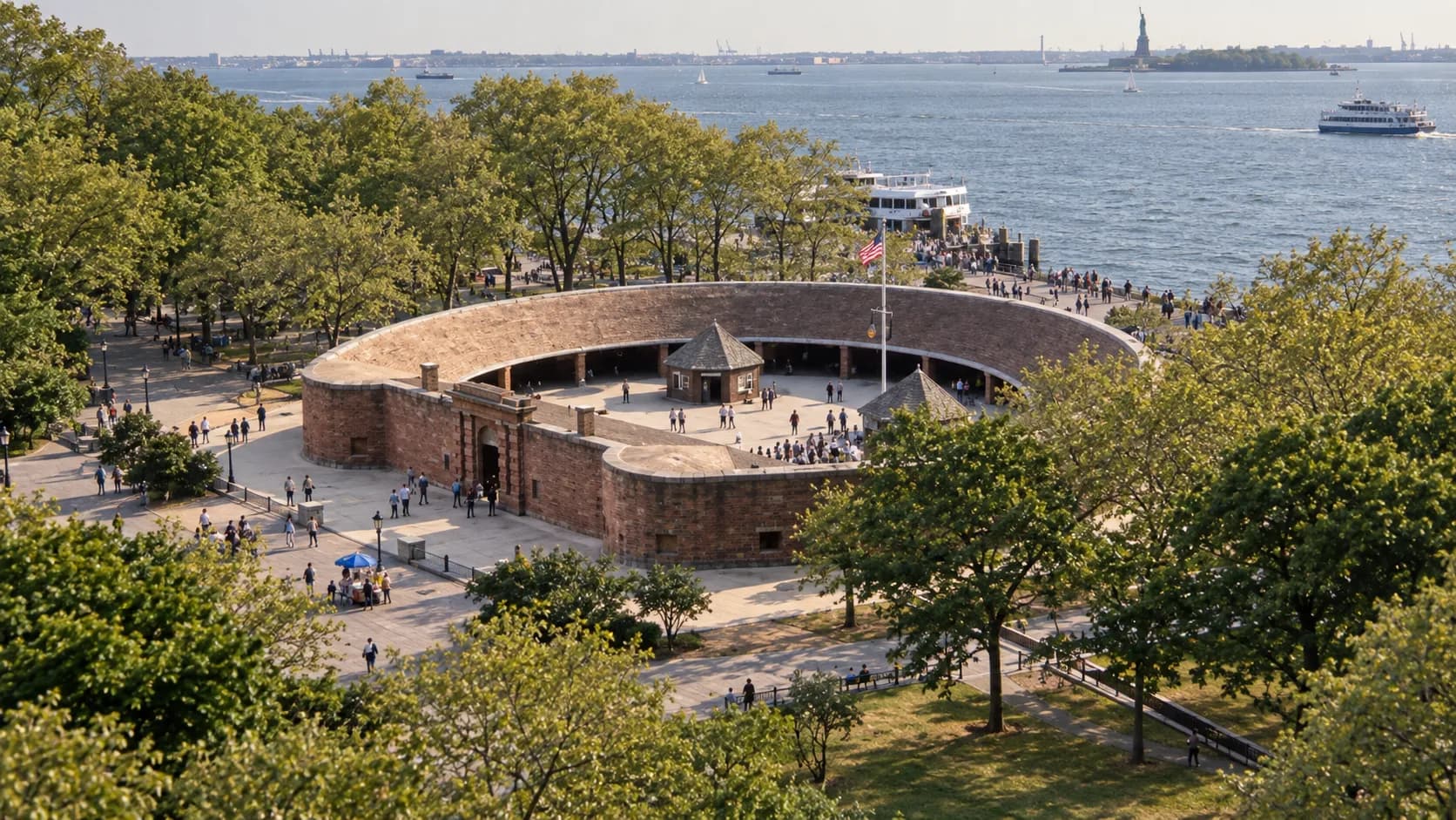Circular reddish-brown Castle Clinton fort in Battery Park, with low masonry walls, open courtyard and blue-grey New York Harbor beyond.
