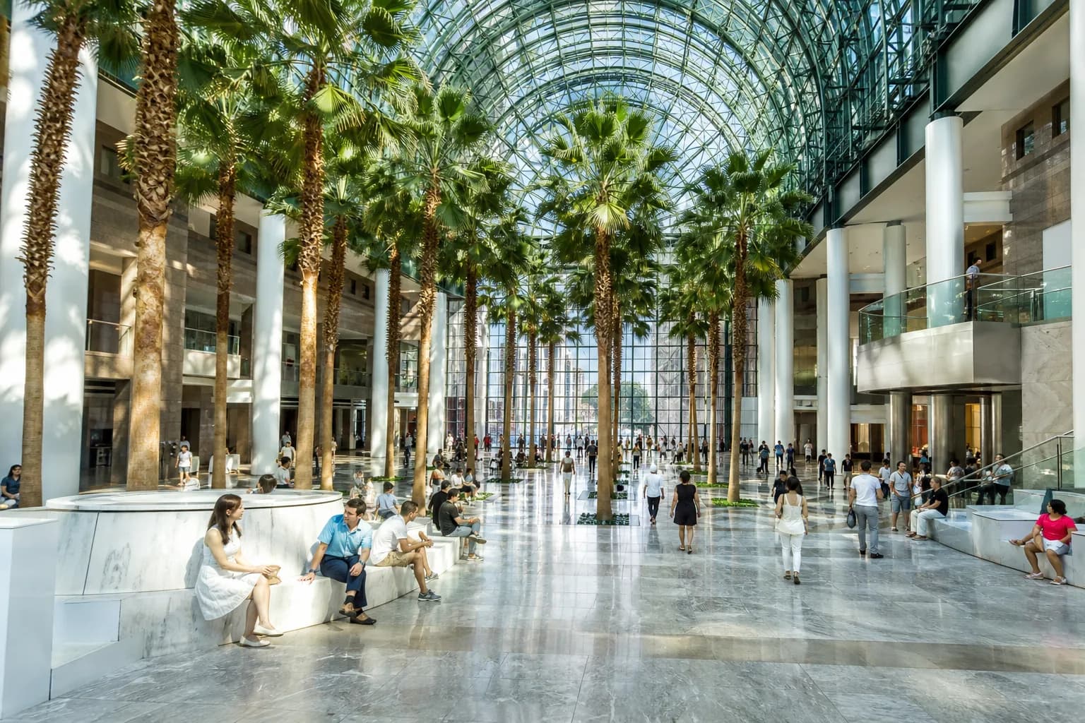 A bright Lower Manhattan indoor public concourse with glass roof, seating, and summer foot traffic