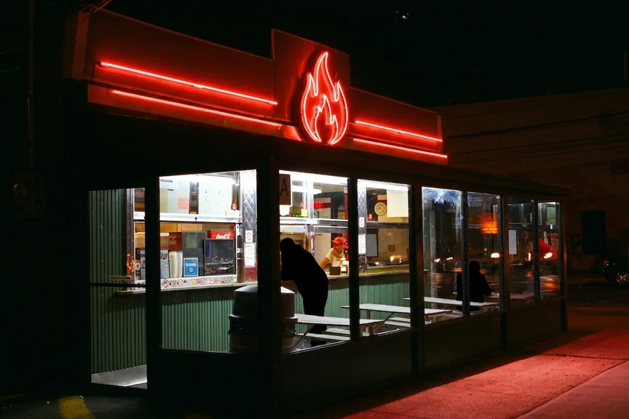 Glass-walled corner pizzeria glowing under a red neon flame sign at night in Howard Beach, Queens