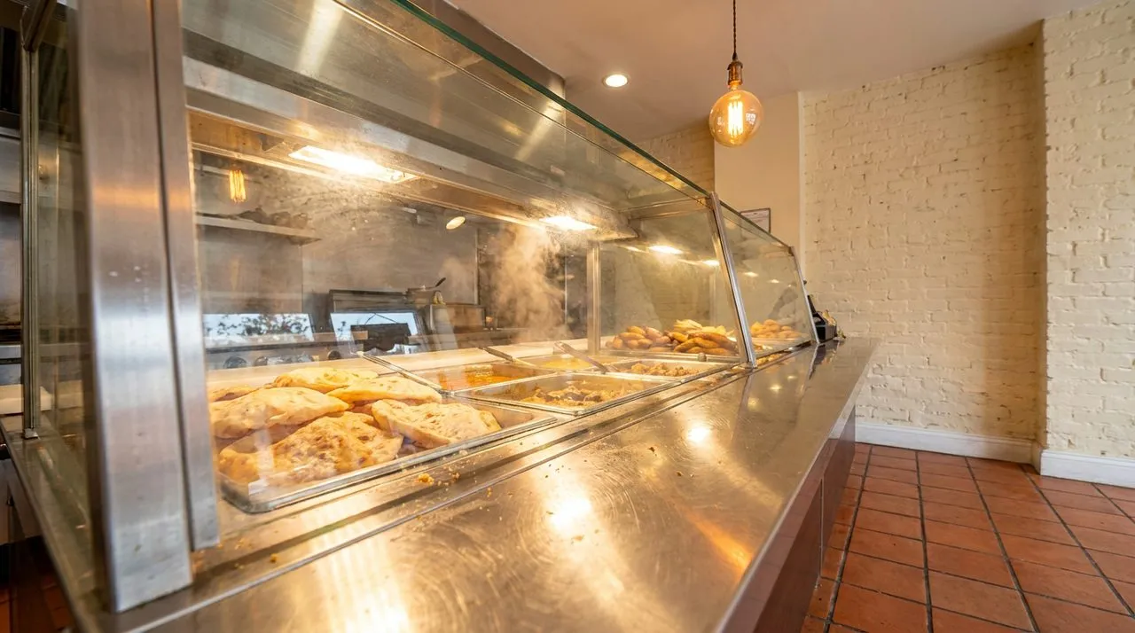 Bright sunny interior of a Crown Heights Trinidadian roti shop with terracotta tiled floor, exposed-brick walls painted creamy white, polished stainless steel counter, glass display case, hanging Edis