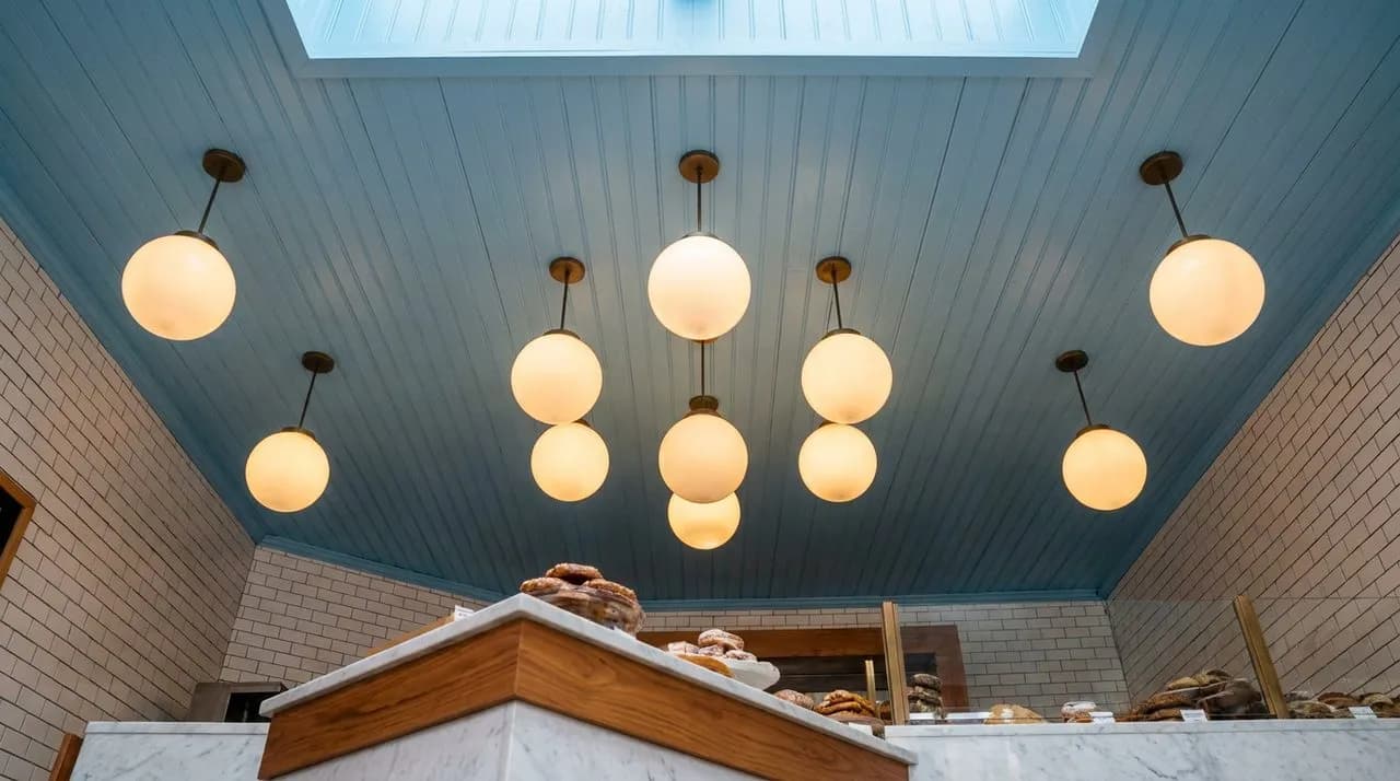 Bright daytime interior of a Brooklyn Haitian bakery with sky-blue painted wood paneling, glass pastry case full of golden patties, white subway-tile walls, warm-colored hanging lights, marble counter