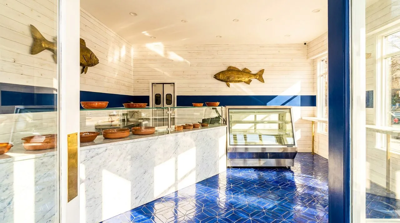 Bright sunny interior of an Astoria Croatian seafood counter with white-washed wood-plank walls, navy-blue accent stripe, polished marble counter, brass fish-shaped wall art, sunlit cobalt tile floor,