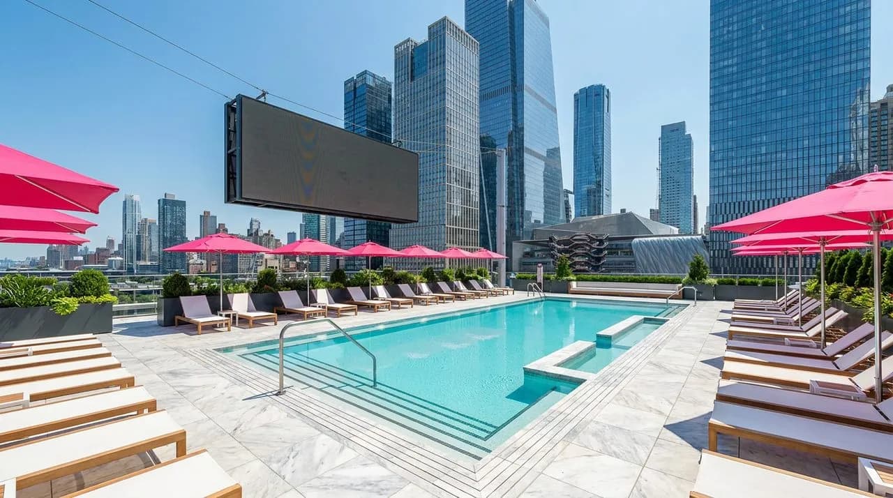 Bright midday view of a Hudson Yards hotel rooftop pool deck with crisp turquoise water, white-and-teak loungers, hot-pink umbrellas, polished marble pool deck, dramatic Manhattan high-rise skyline, l