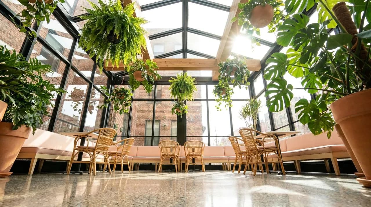 Bright midday view of a Brooklyn boutique-hotel solarium with floor-to-ceiling glass walls, hanging tropical plants, rattan chairs, pale-pink cushioned banquettes, polished terrazzo floor, sky-bright 