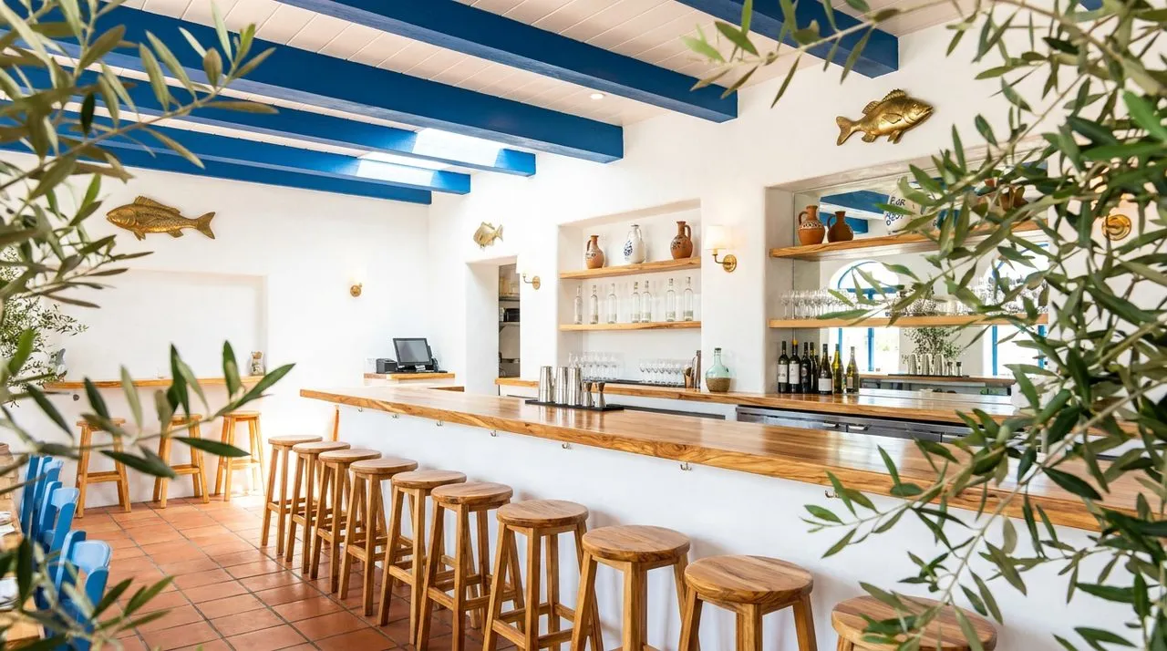 Bright sunny daytime view of an Astoria Greek taverna interior with whitewashed plaster walls, deep-blue painted wood ceiling beams, terracotta tile floor, polished olive-wood counter, brass fish wall