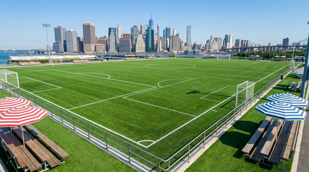 Bright sunny afternoon view of Pier 5 Brooklyn Bridge Park multipurpose field, vivid green turf, polished steel railings, Manhattan skyline backdrop, vintage wooden bleacher seating, large red-and-whi