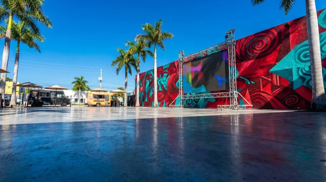 Bright sunny afternoon view of a Wynwood open-air plaza with vivid hand-painted mural wall in saturated reds, oranges, and turquoise (abstract geometric patterns, no readable text), polished concrete 