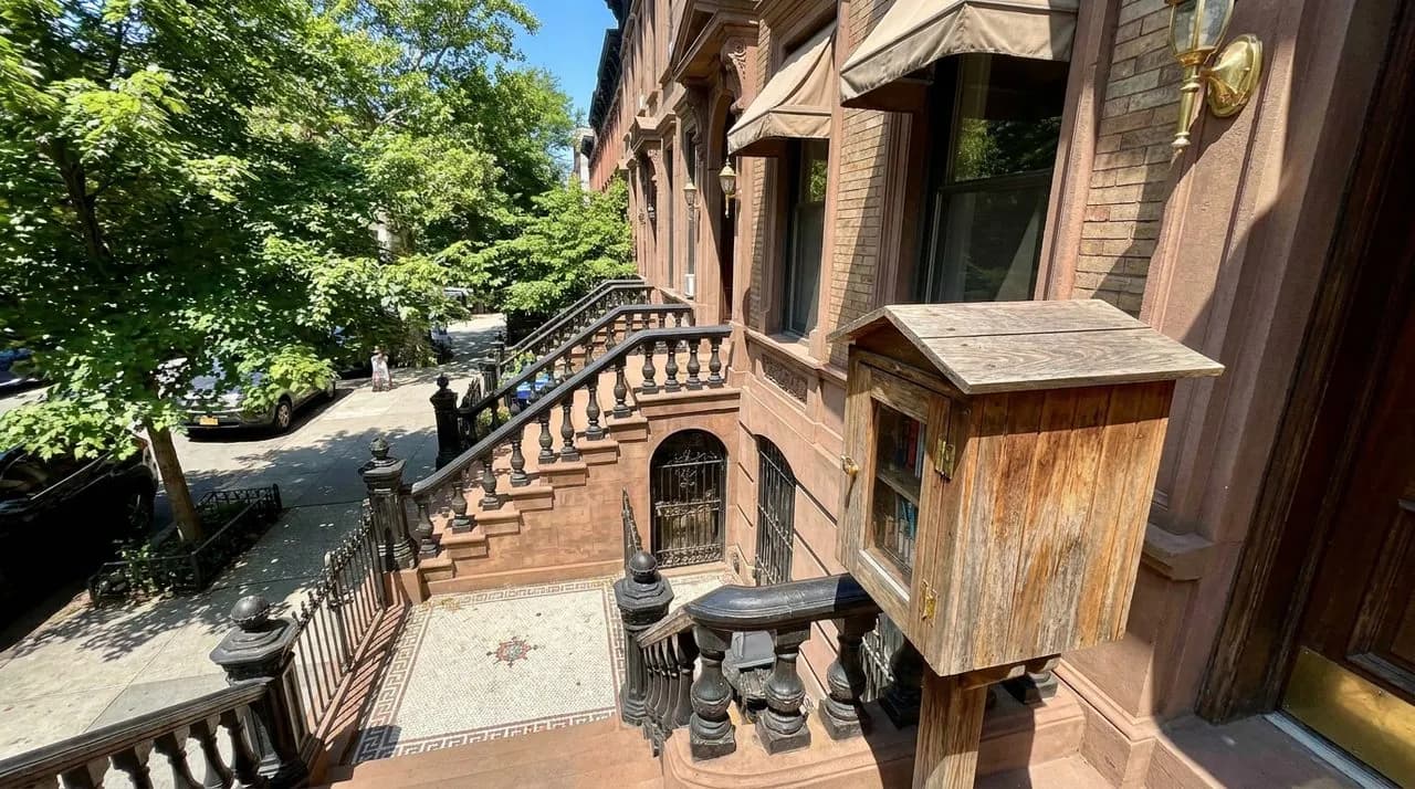 Bright sunny daytime wide-angle of a Brooklyn brownstone street with a row of stoops and a small wooden lending box at corner, leafy summer canopy, vivid blue sky, polished brownstone steps. No people