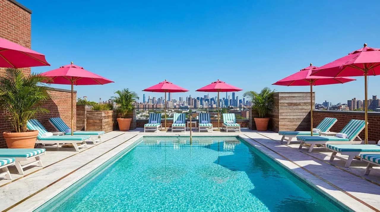Bright sunny afternoon Williamsburg hotel rooftop pool deck with turquoise sparkling water, crisp white teak loungers, hot-pink umbrellas, polished marble decking, lush potted palms, panoramic Manhatt