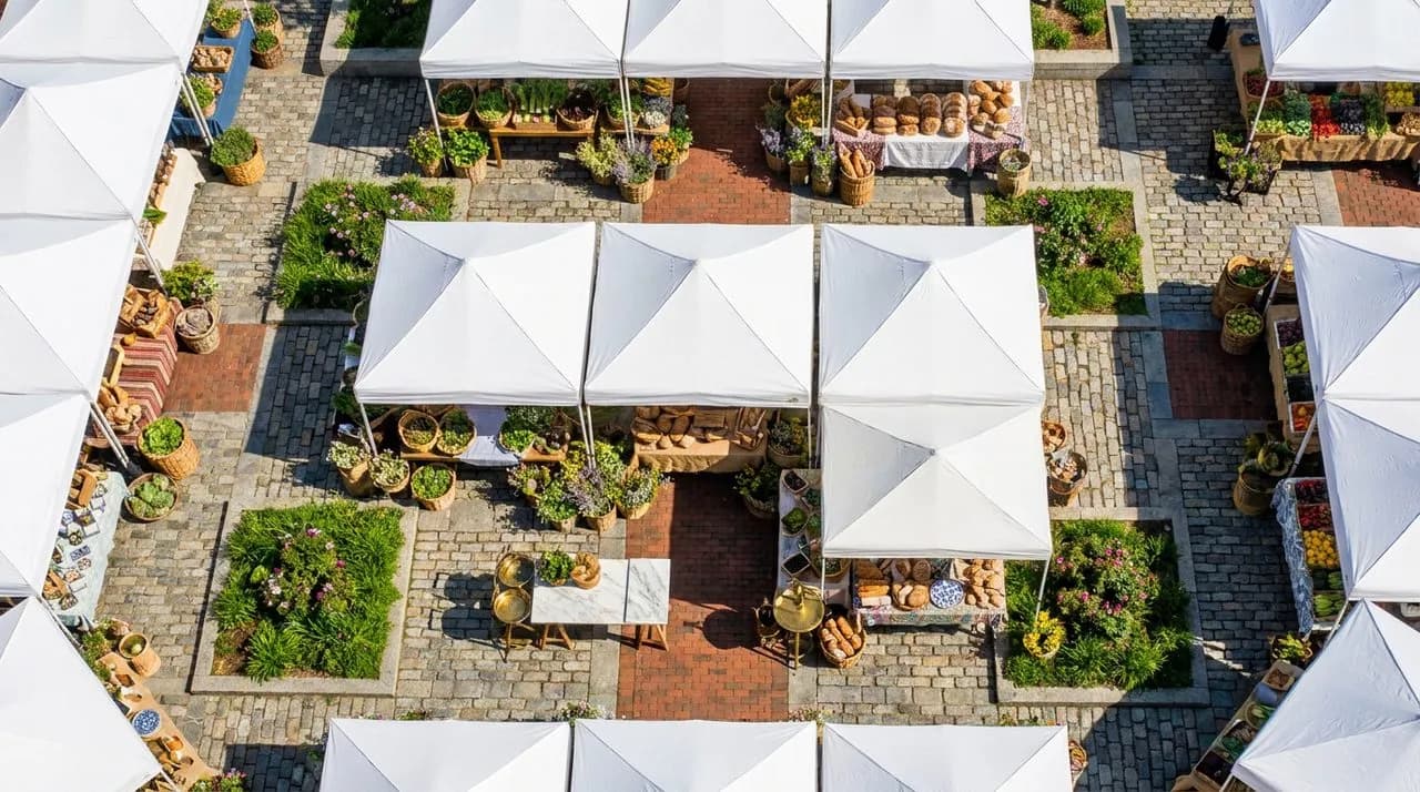 Bright sunny day high-angle overhead of a Grand Army Plaza farmers market layout with white tent stalls in grid, baskets of spring greens and flowers, polished cobblestone plaza, vivid blue sky. No pe