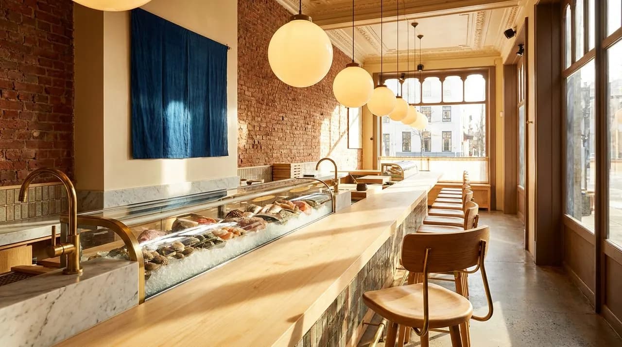 Bright sunny early-evening interior of an East Village omakase counter with blonde hinoki wood bar, indigo-blue cloth banner on cream wall, single empty wood stool foreground, polished glass display o
