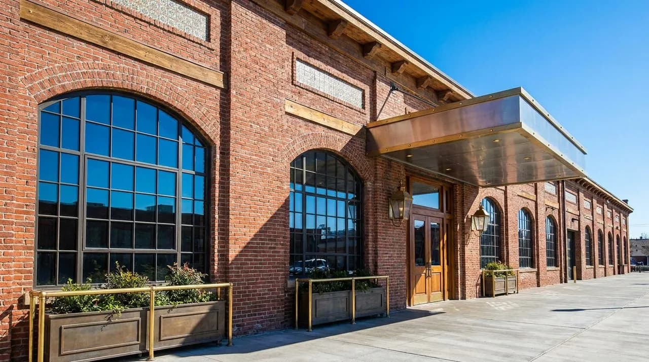 Bright daytime exterior of a Randolph Row converted warehouse facade with weathered red brick, large industrial windows, brushed steel awning, vivid blue sky, polished concrete sidewalk. No people. Ph