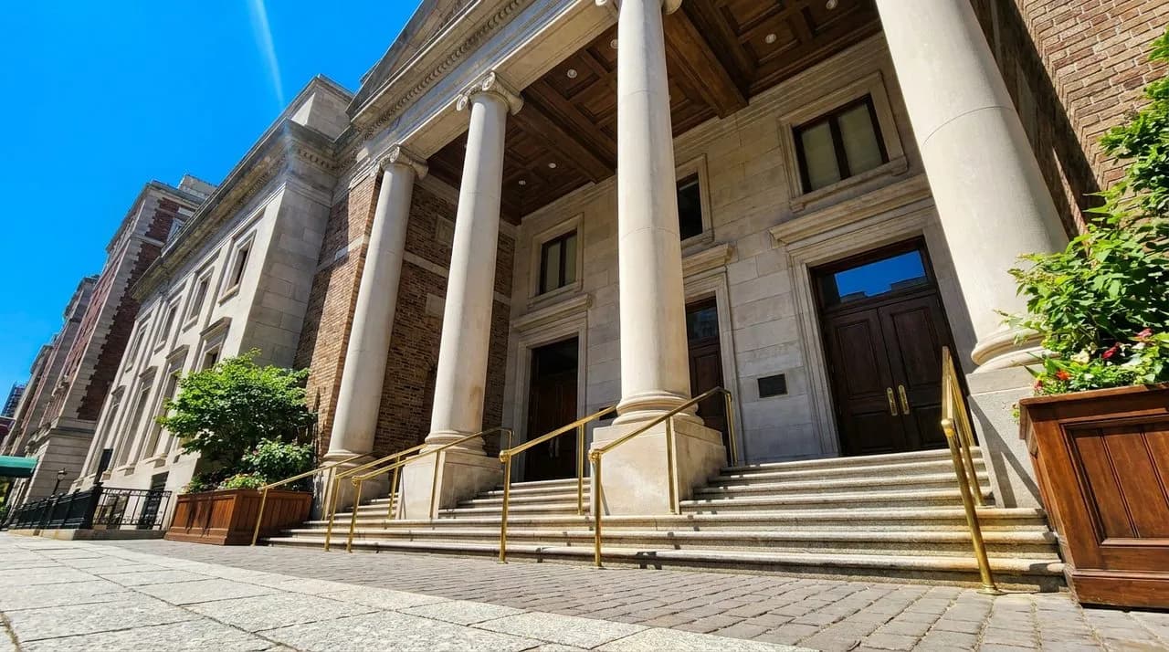 Bright sunny daytime exterior of a grand NYC museum facade with limestone columns, wide stone steps, polished brass railings, vivid blue sky, leafy summer trees framing. No people. Photo-realistic edi