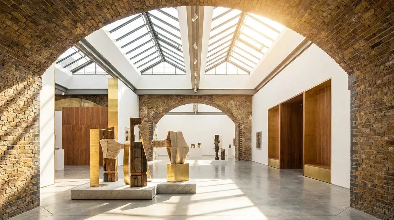 Bright daytime interior of a contemporary museum gallery with polished concrete floor, white walls, large arched skylights flooding with natural light, abstract sculpture forms in foreground (generic,