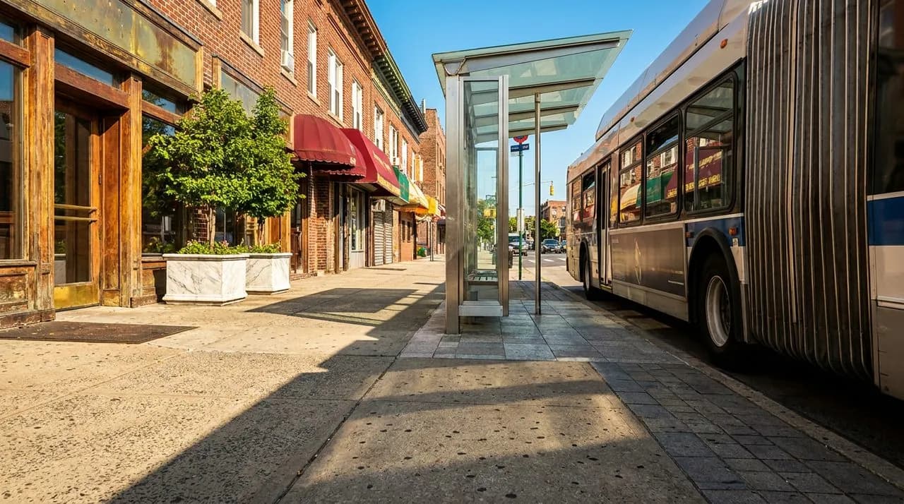 Bright daytime exterior of an MTA city bus pulling up to a Queens neighborhood stop with polished glass bus shelter, weathered red-brick storefronts, leafy summer trees, vivid blue sky, polished concr
