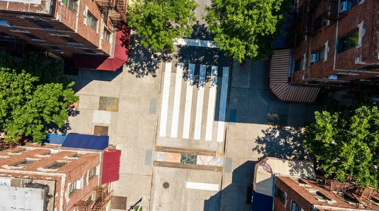 Bright daytime overhead view of a Bronx pedestrian crosswalk with polished concrete, painted white stripes, leafy summer trees, vivid blue sky, weathered brick apartment building backdrop. No people. 