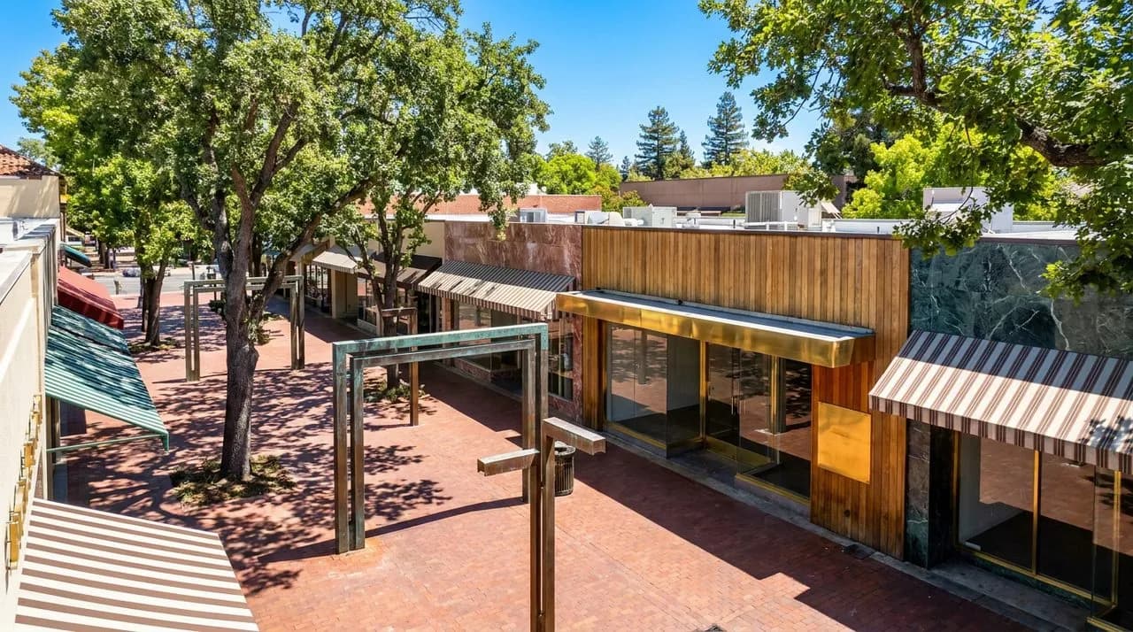 Bright sunny day Palo Alto University Avenue view with leafy oak trees, polished red-brick sidewalk, mid-century modern storefronts, vivid blue sky, weathered bronze street fixtures. No people. Photo-