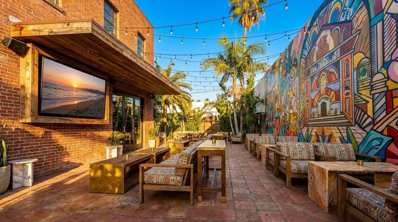 Bright sunny LA late-afternoon Echo Park lounge patio, colorful mural wall on one side, large outdoor TV mounted under awning, string lights, terracotta tile floor, palm trees, vivid blue sky