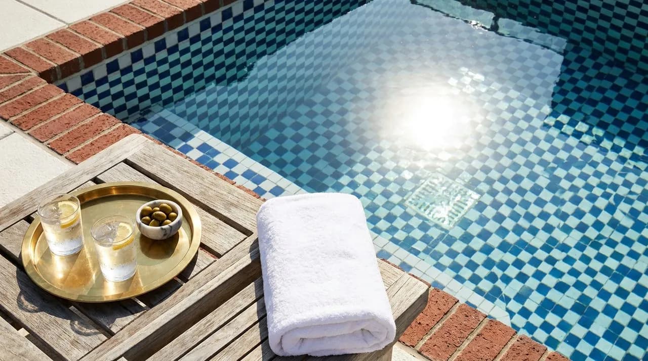Bright sunny noon overhead detail of a NYC rooftop pool corner, patterned blue-and-white tile, neatly folded white towel, brass drinks tray, sun reflection on water