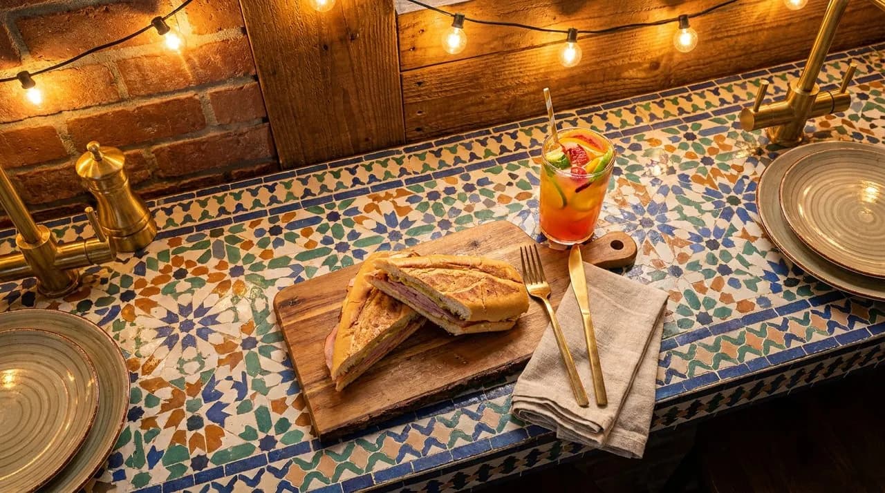Bright Miami early-evening overhead detail of a Wynwood food-truck table, patterned tile surface, freshly pressed Cuban sandwich, colorful tropical drink, brass utensils, warm string-light glow