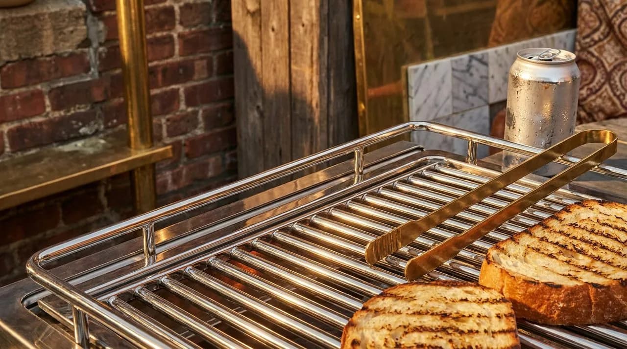 Extreme close-up of a chrome backyard BBQ grill at a Brooklyn block party, brass tongs resting on rail, freshly grilled bread with crisp grill marks, condensation on a drink can, soft golden raking li