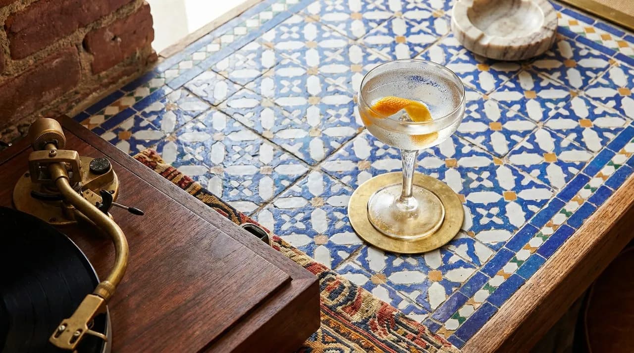 Bright sunny midday overhead detail of a tile-inlaid counter at a LES listening bar, single coupe cocktail with citrus twist, tip of a turntable tonearm visible at edge, brass coaster, vivid pattern
