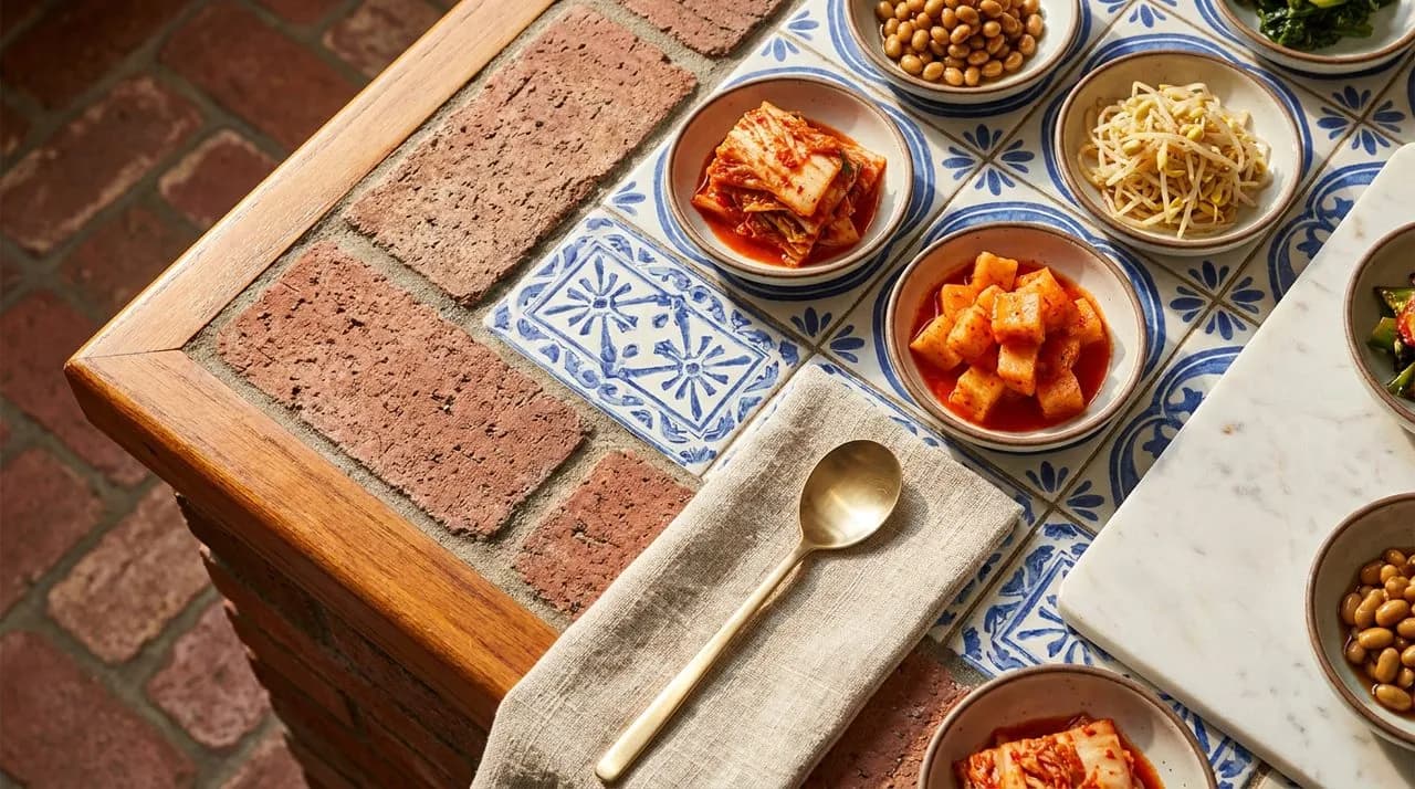 Bright sunny midday overhead detail of a Koreatown counter side-dish setup, patterned tile inset under small ceramic banchan dishes, kimchi color contrast, brass spoon on cloth, polished wood edge