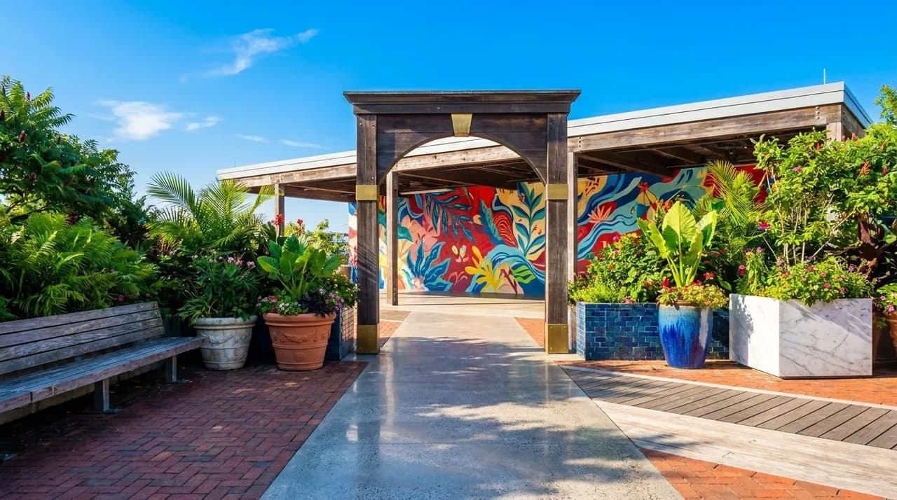 Bright sunny morning view of a Brooklyn Bridge Park yoga pavilion, colorful mural backdrop on far wall, leafy planters, polished concrete path, vivid blue sky, weathered wood bench