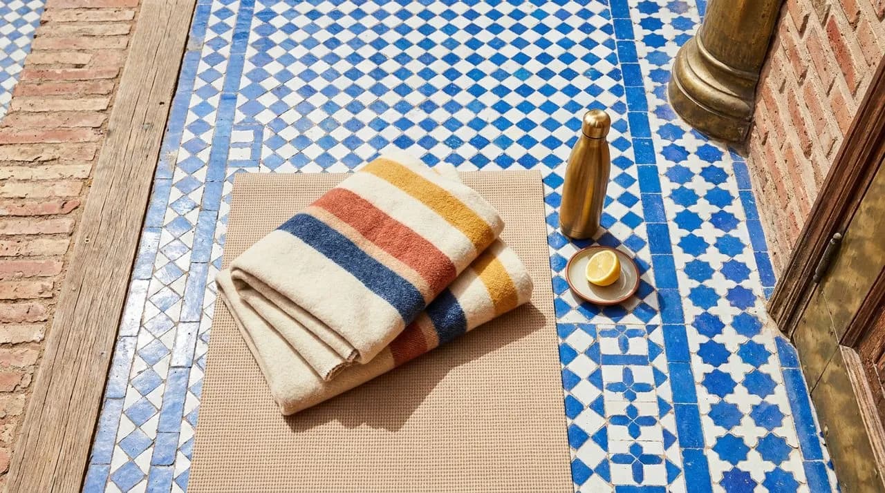 Bright sunny midday overhead detail of a yoga mat on a tile path inset, neatly folded wool blanket, brass insulated water bottle, fresh lemon wedge on a saucer, vivid pattern color contrast