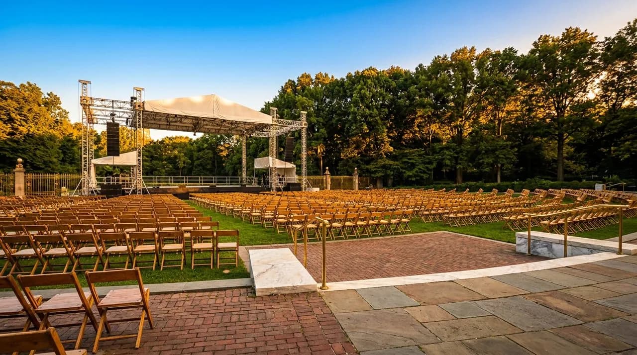 Bright sunny late-afternoon Central Park SummerStage lawn at Rumsey Playfield, stage scaffolding with white canopy, tall tree line backdrop, folding chairs neatly arranged on grass, vivid sky, no peop