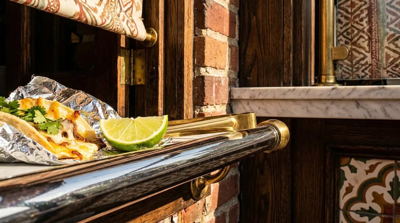 Extreme close-up of a Brooklyn taqueria service window chrome rail, foil-wrapped taco edge, fresh lime wedge, brass tongs resting on rail, side raking afternoon light, vivid color