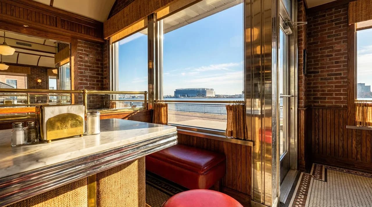 Bright sunny day view through chrome NYC diner window framing distant MetLife Stadium silhouette across river, polished chrome counter foreground, brass napkin holder, vinyl booth, soft daylight