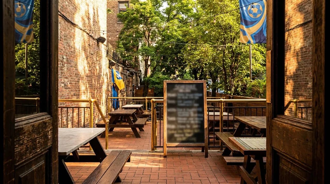 Bright sunny late-afternoon NYC bar patio set for WC 2026 watch party, picnic tables, chalkboard A-frame schedule, string lights, leafy summer trees, vivid golden light, polished metal railings