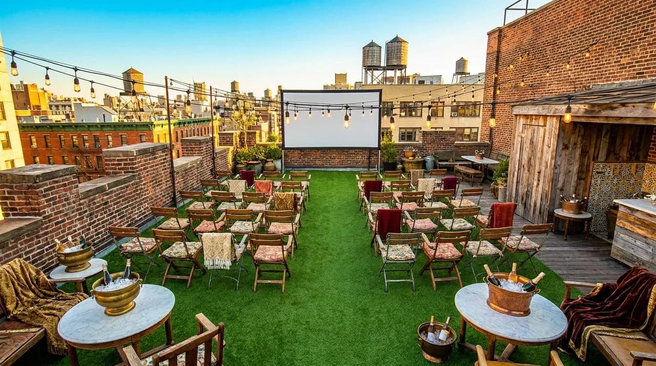 Bright sunny golden-hour overhead view of NYC rooftop lawn party setup for WC 2026 final, lawn chairs in rows, large projector screen, festoon lights, ice buckets, no people, vivid sky