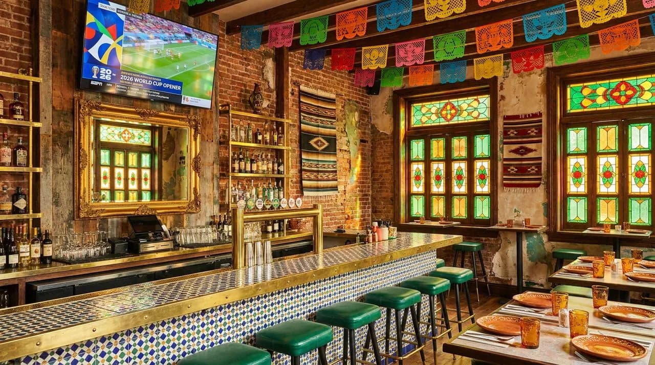 Bright sunny afternoon Jackson Heights Mexican cantina interior set for WC 2026 opener, patterned tile counter, emerald green accents, brass-framed mirror, papel picado overhead, large TV, no people