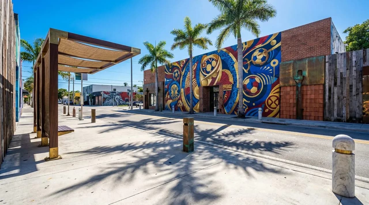 Bright sunny Miami midday wide view of Wynwood mural street on WC 2026 match day, saturated colorful mural, palm tree shadows, polished concrete sidewalk, vivid blue sky, bus stop in frame, no people