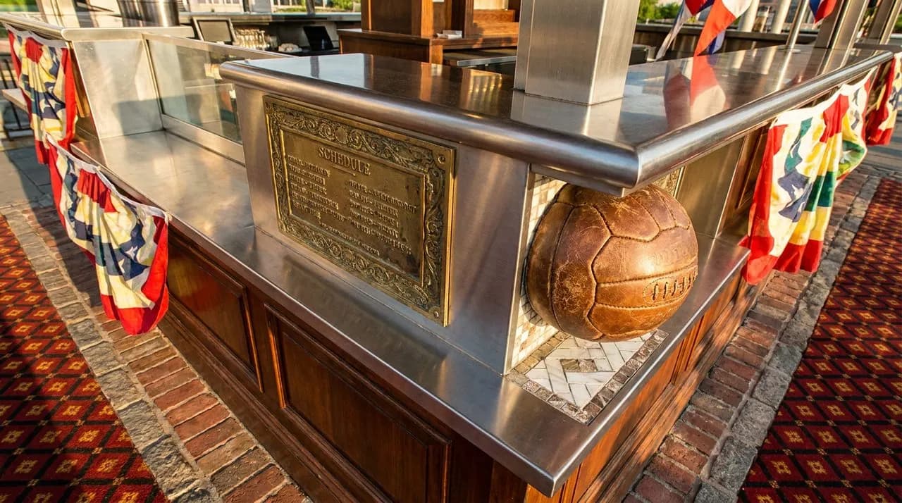 Extreme close-up of Brooklyn Bridge Park festival kiosk for WC 2026 fan festival, polished steel kiosk, brass schedule plaque, soccer ball decorative detail, warm afternoon light