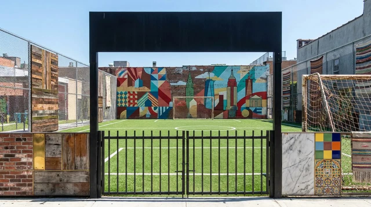 Bright sunny midday wide view of free NYC public soccer pitch for WC 2026 pickup, green turf, chain-link fence, mural backdrop wall, weathered metal goals, painted sideline, vivid blue sky, no people