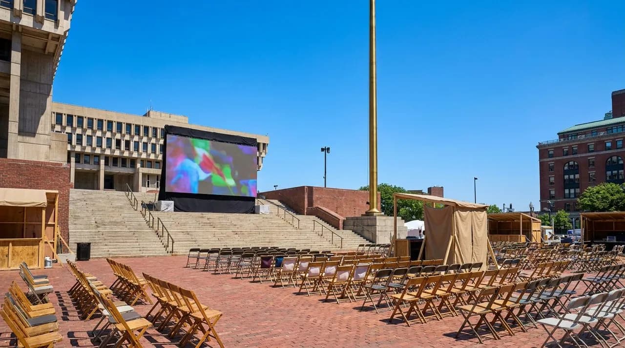 Bright sunny midday wide view of Boston City Hall Plaza set for WC 2026 free watch party, brick plaza, concrete steps, large projection screen, brass flagpole, vivid blue sky, no people