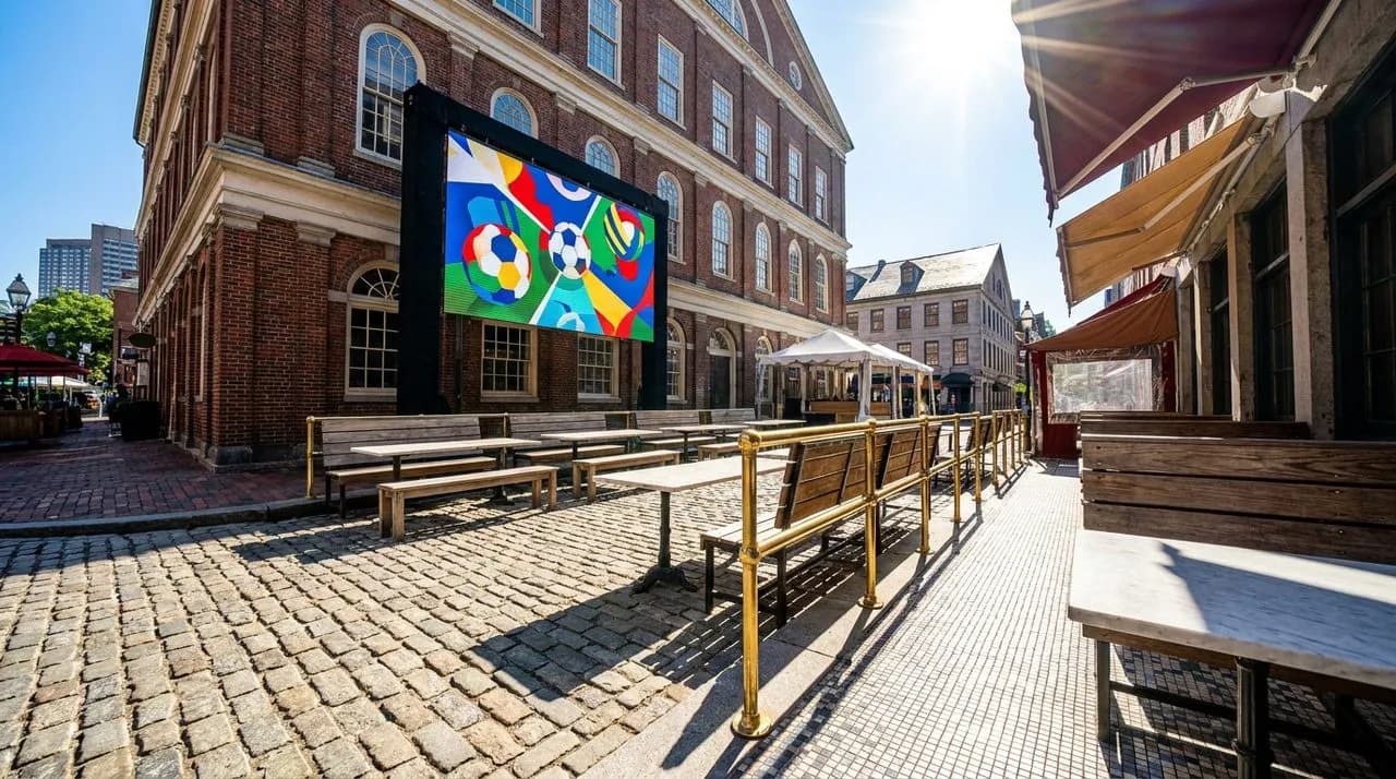 Bright sunny midday low-angle Faneuil Hall outdoor area set for WC 2026 free watch party, cobblestone street, brick facade, large outdoor screen, brass railings, no people